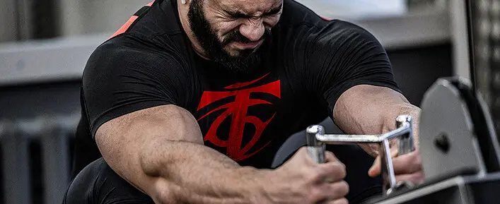 A Muscular Man In A Black Shirt With A Red Design Is Intensely Working Out On A Gym Machine, Focusing On Muscle Growth And Strength Training, Showcasing The Effects Of Deca Steroids On Muscle Development.